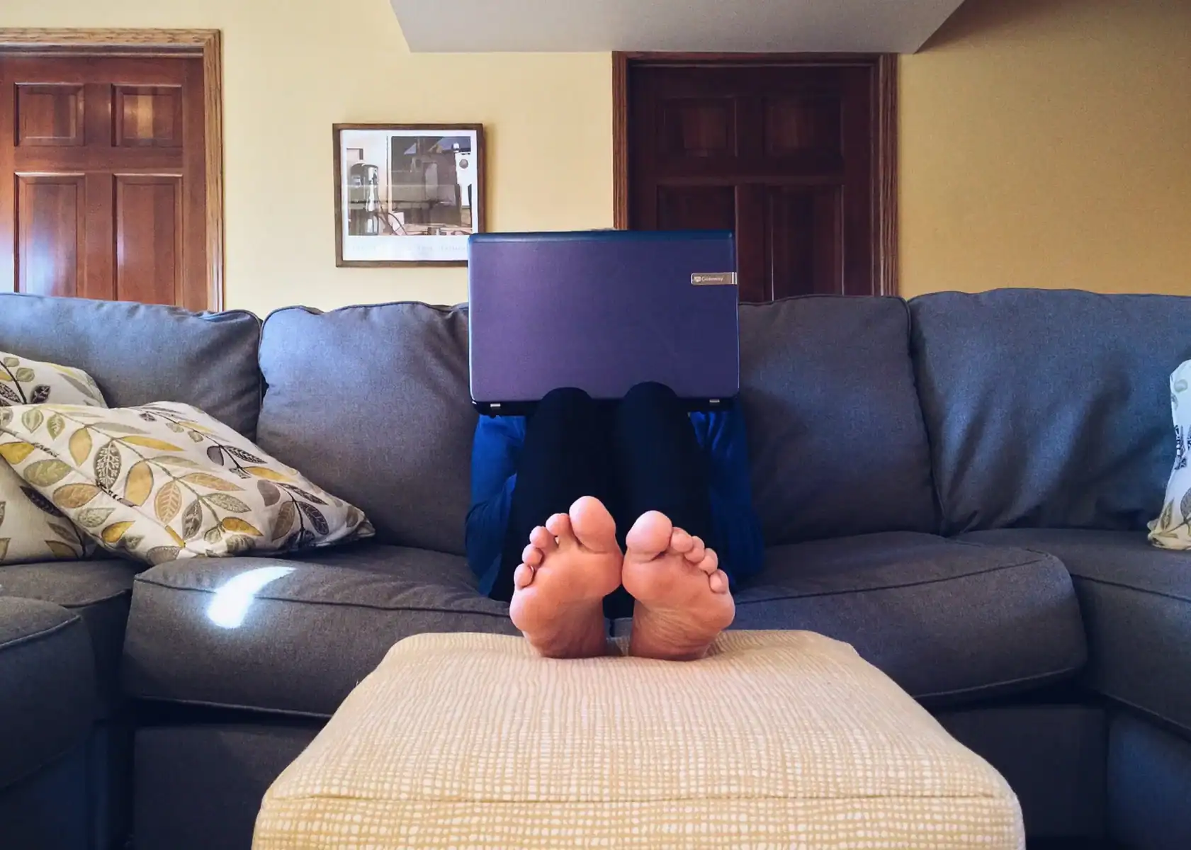 Person sitting on a gray couch with their feet up on a beige ottoman, using a laptop that hides their face. The background shows wooden doors and a framed picture on the wall, hinting at the balance of the Battle of Work and Family in an empowered relationship.