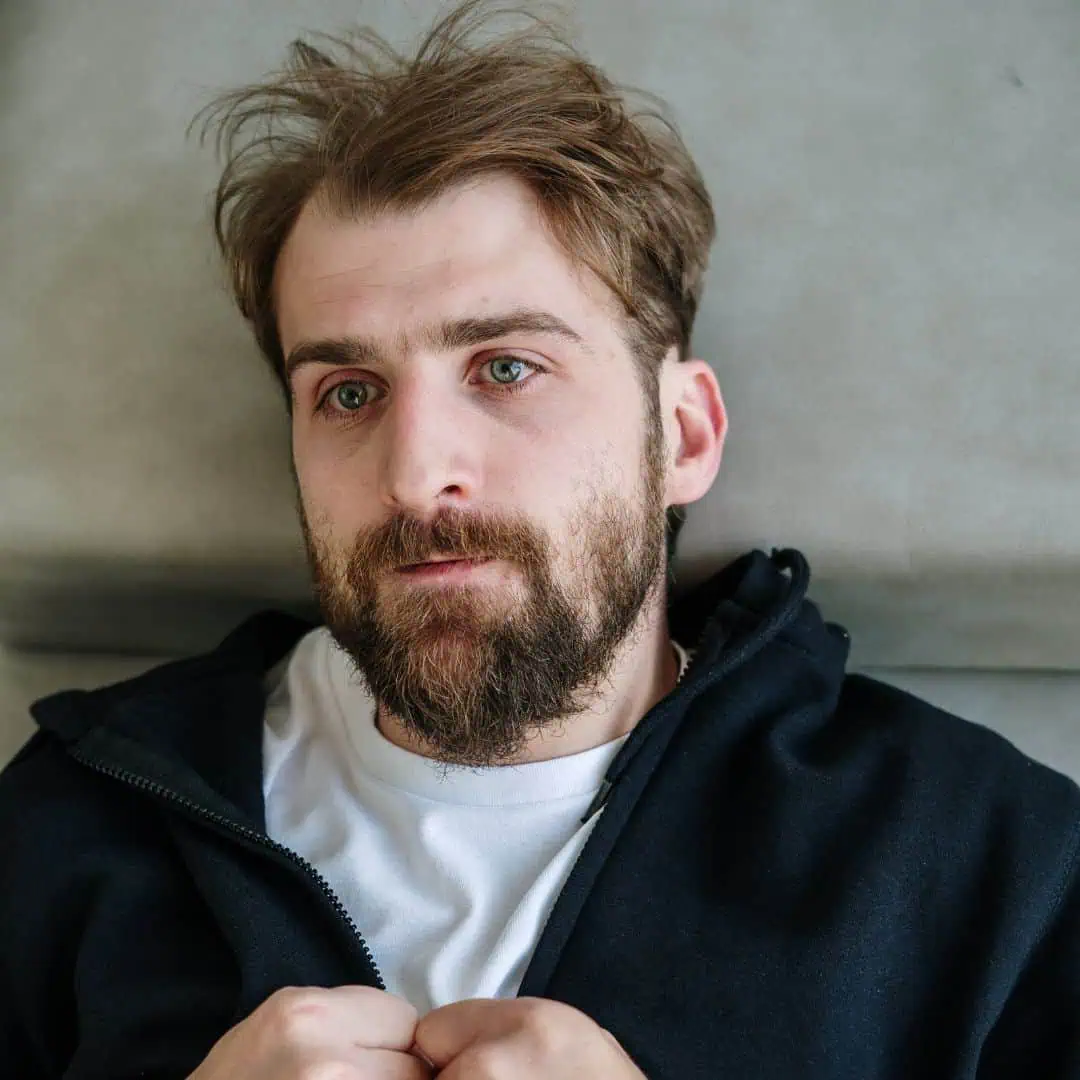 A bearded man with light brown hair, slightly messy, is lying on a gray surface. He is wearing a dark zip-up hoodie over a white t-shirt, and his hands are loosely holding the edges of the hoodie. Gazing off into the distance with a contemplative expression, he reflects on men's mental health issues.