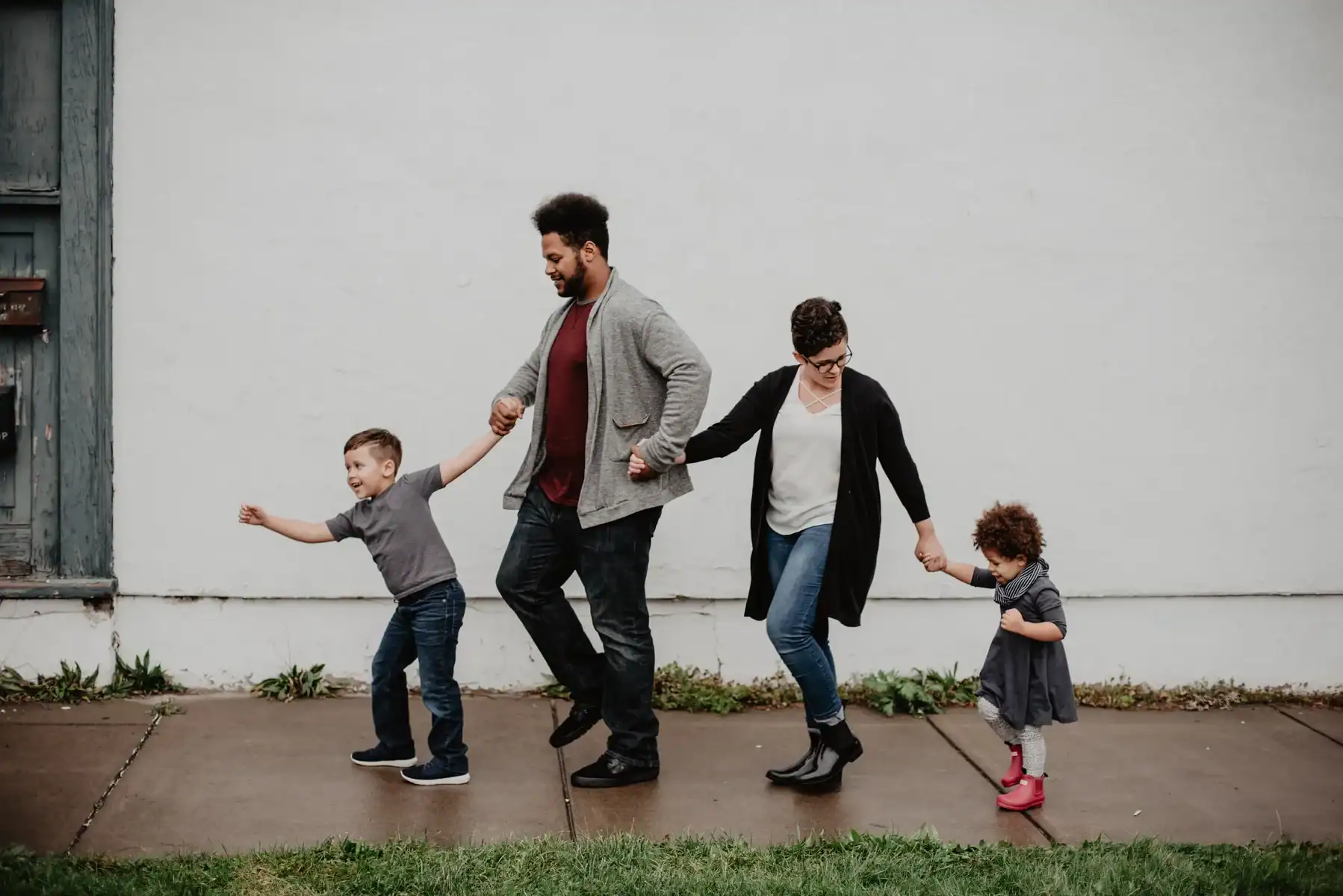 A family of four walks hand in hand on a sidewalk in front of a white wall. The father and mother are in the middle, holding the hands of their young son on the left and their young daughter on the right. Everyone is smiling and appears cheerful, embodying tips and tricks for a happy marriage.