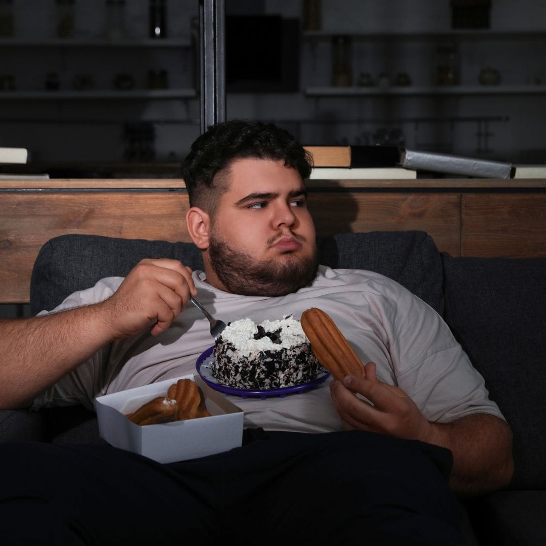A person with short dark hair, wearing a light gray t-shirt, sits on a couch holding a partially eaten chocolate cake with whipped cream on a plate, a fork in one hand, and a churro in the other. A box with more churros is on their lap. The background is dimly lit, hinting at signs of male depression often ignored.