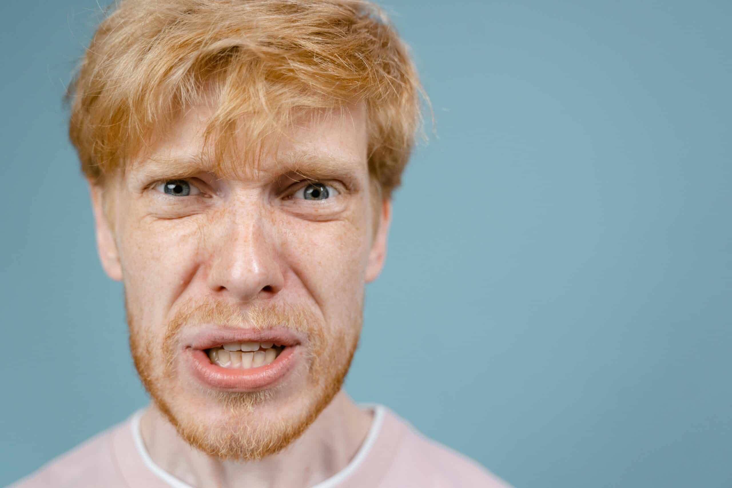 A person with red hair and a beard is looking directly at the camera with a tense expression against a blue background. This emotionally intelligent man seems to be expressing frustration or anger, perhaps reflecting on the complexities of relationships.