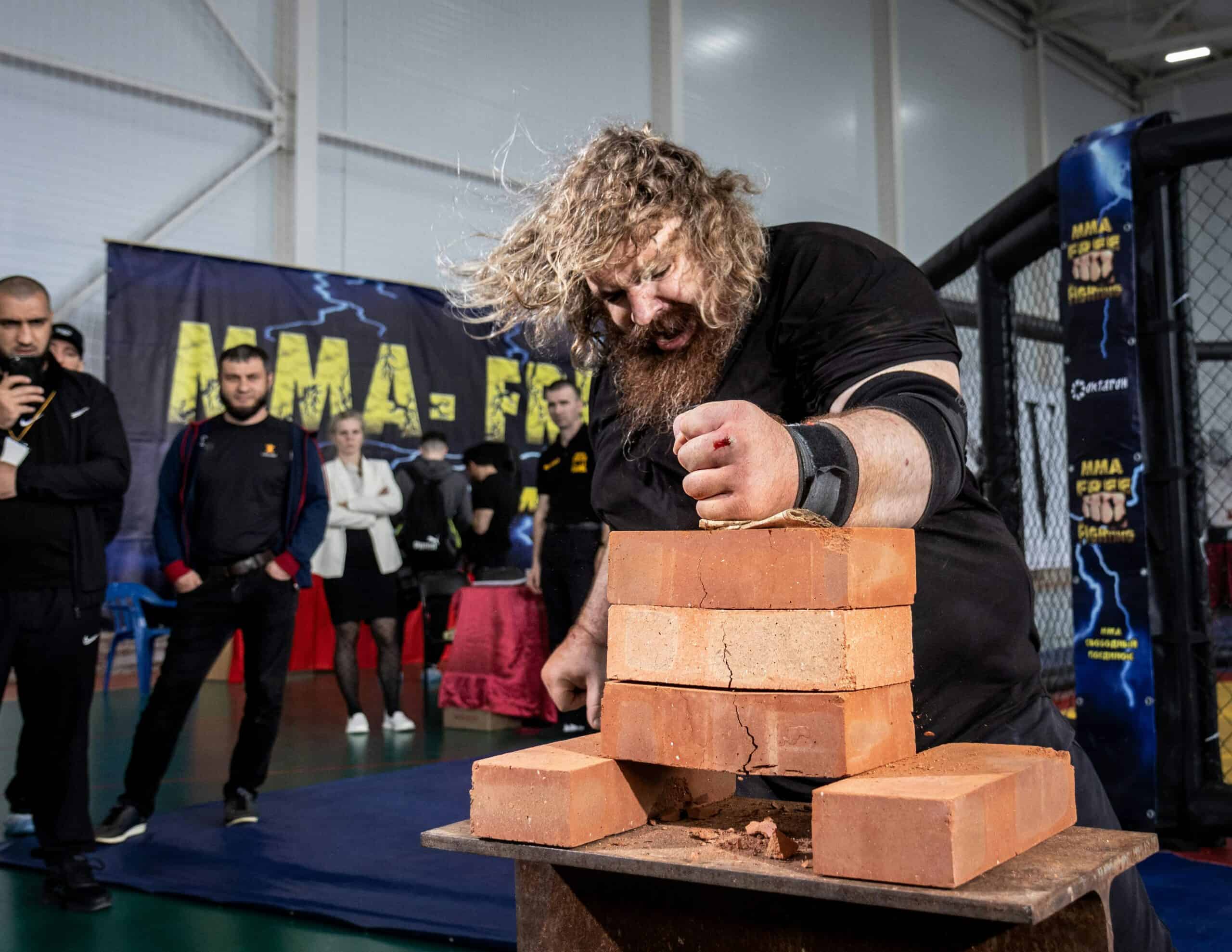 At a martial arts event, a man with long hair and a beard channels unexpected power as he attempts to break a stack of clay bricks with his hand. Spectators and officials stand nearby, observing the action, captivated by this display of strength and vulnerability.