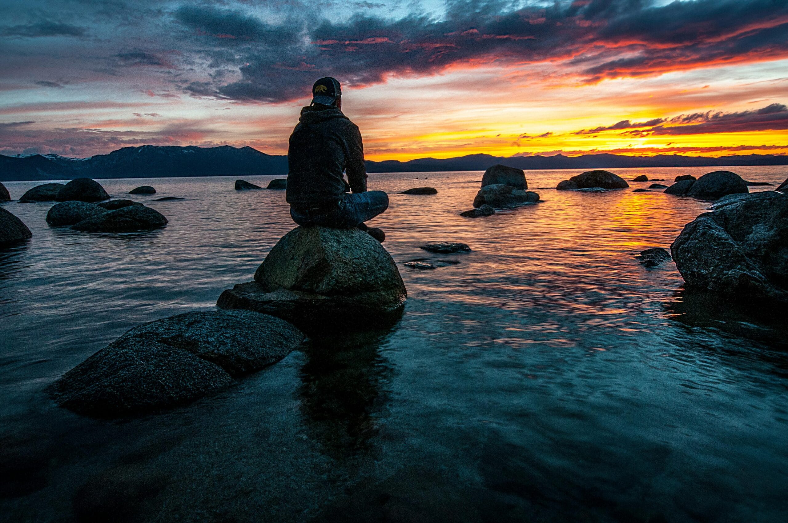 A man sits on a rock by a tranquil lake, watching a vibrant sunset with orange, pink, and purple hues in the sky. The calm water reflects the colorful sky, and distant mountains frame the horizon.