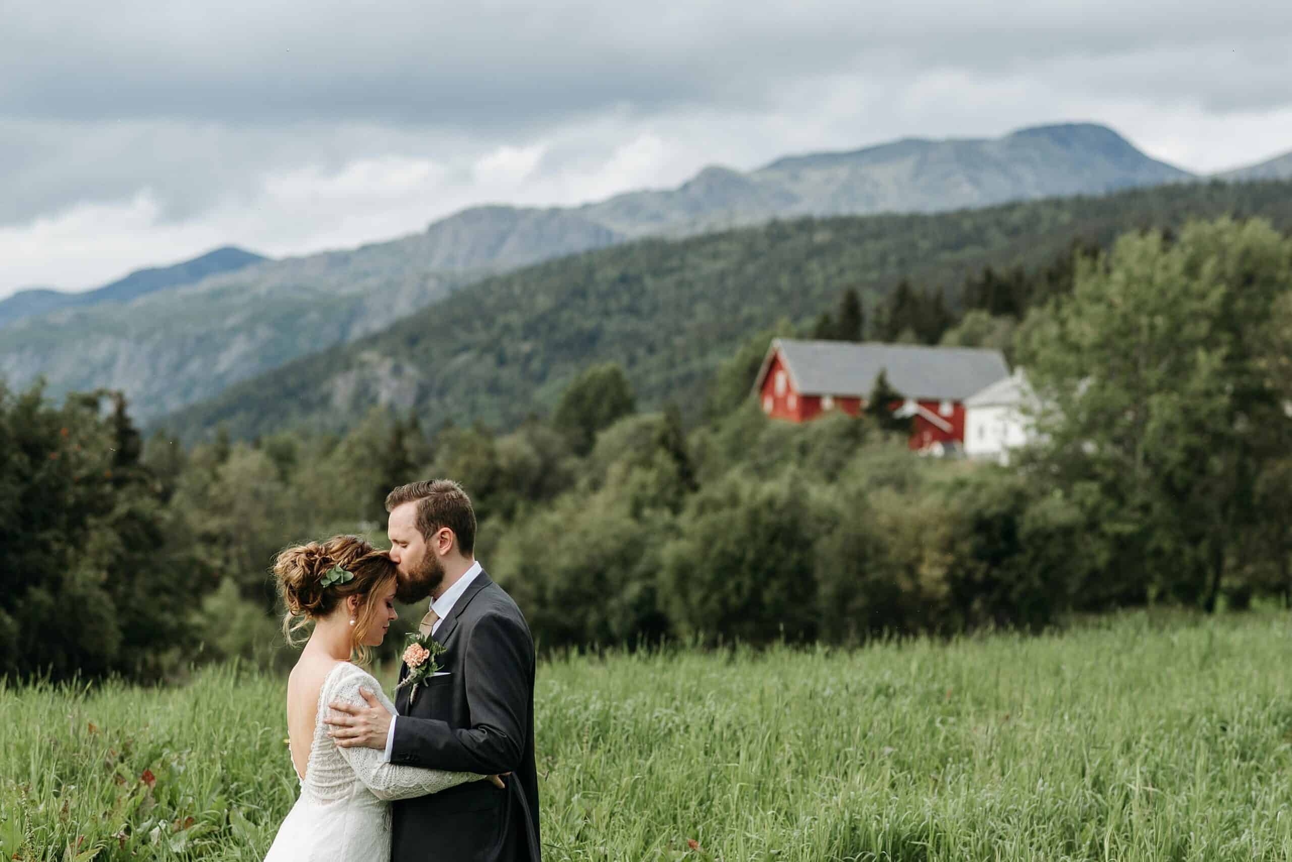 A bride and groom, embodying the essence of a good marriage, embrace in a lush green field with mountains in the background. A red and white house sits among trees under a cloudy sky, symbolizing the foundation of their promising future together.