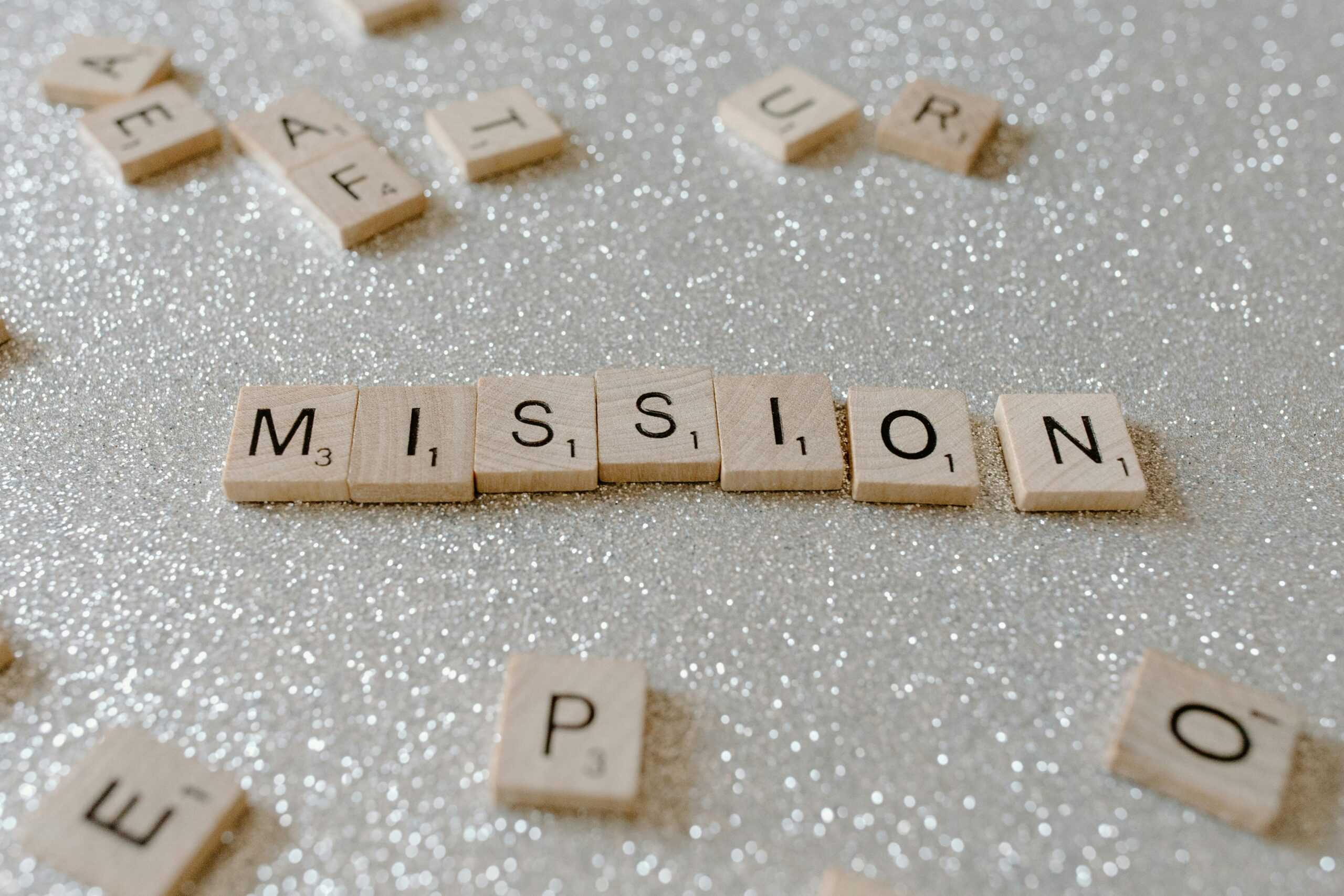 Scrabble tiles spelling "MISSION" are arranged on a sparkling, glittery surface, surrounded by other scattered tiles. It's as if they whisper: Stop Setting Goals like men setting sparks—start setting fires to ignite your true purpose.