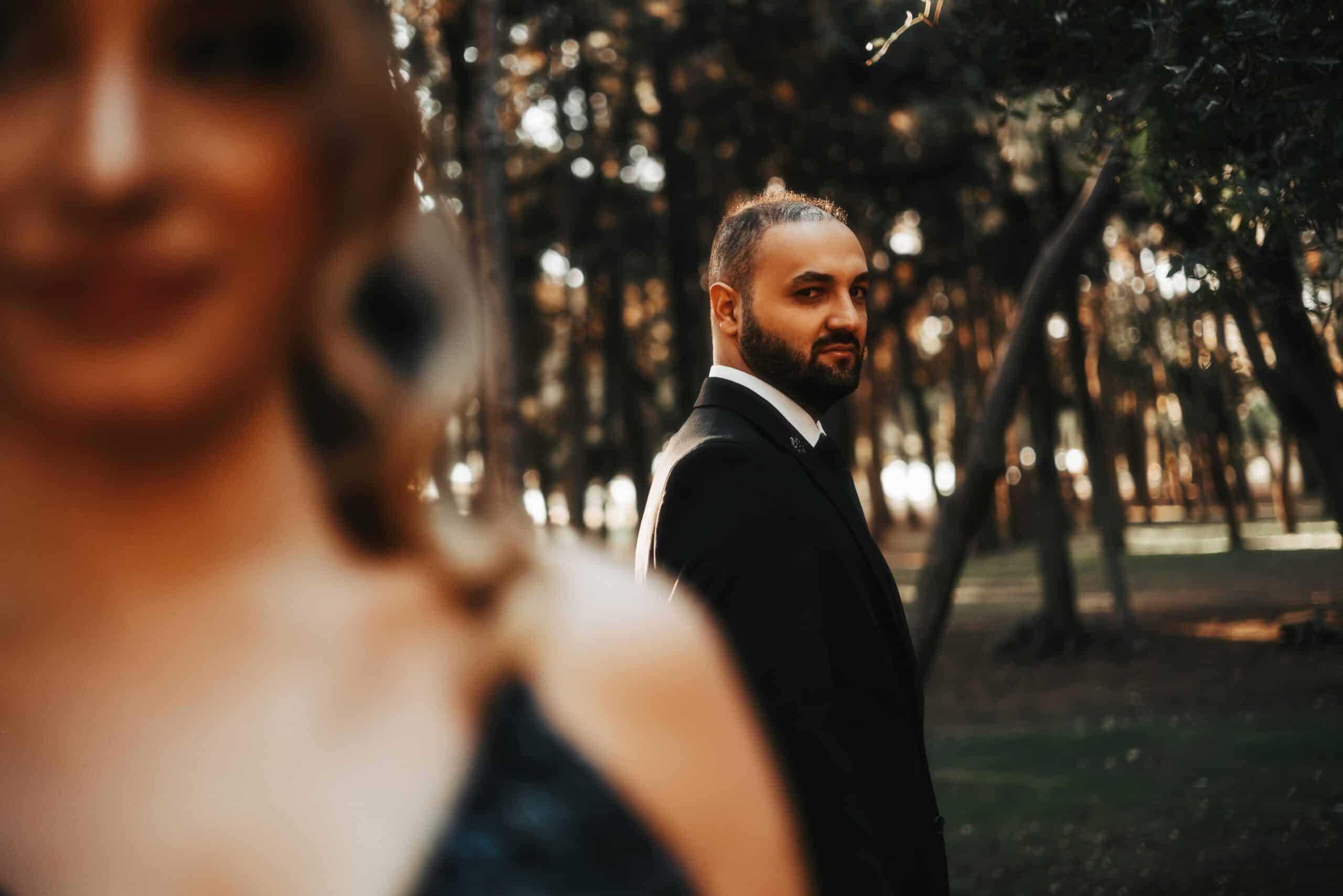 A man in a suit stands in focus against a blurred forest background, seemingly avoiding conversation. In the foreground, the blurry outline of a woman's shoulder and hair is visible. The scene has a warm, golden glow, hinting at an important relationship discussion just out of reach.